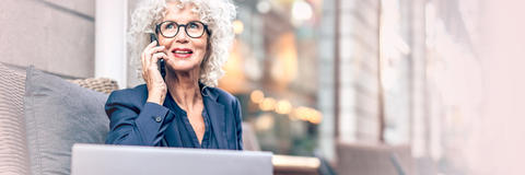 Frau sitzt vor ihrem Laptop und telefoniert mit ihrem Smartphone