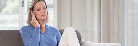 Eine Frau sitzt mit geschlossenen Augen auf einem Sofa und hält sich wegen Ohrgeräuschen die Hand ans Ohr