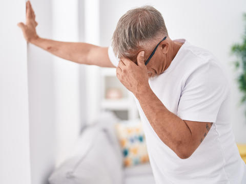 Eine Mann greift sich wegen Schwindel mit der einen Hand an den Kopf und stützt sich mit der anderen Hand an der Wand ab
