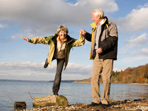 Eine ältere Dame balanciert bei einem Spaziergang auf einem Stein. Ihr Partner hält sie dabei an der Hand fest.